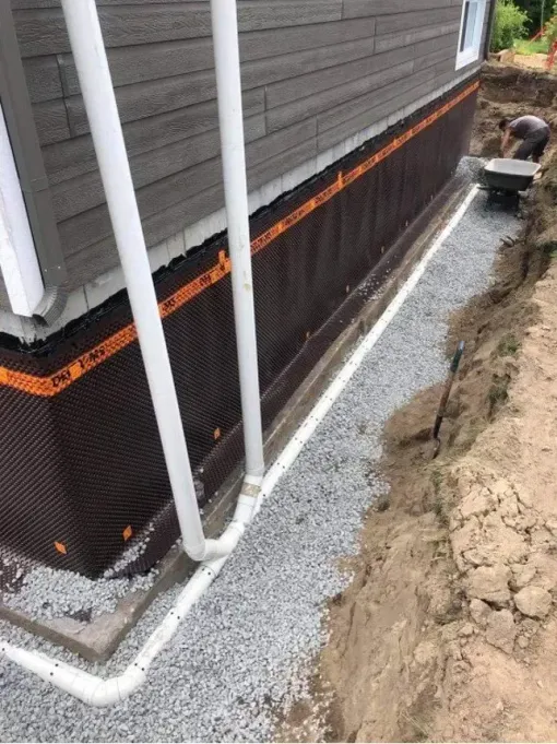 House foundation wrapped in black waterproofing and orange trim beside a gravel trench and pipes