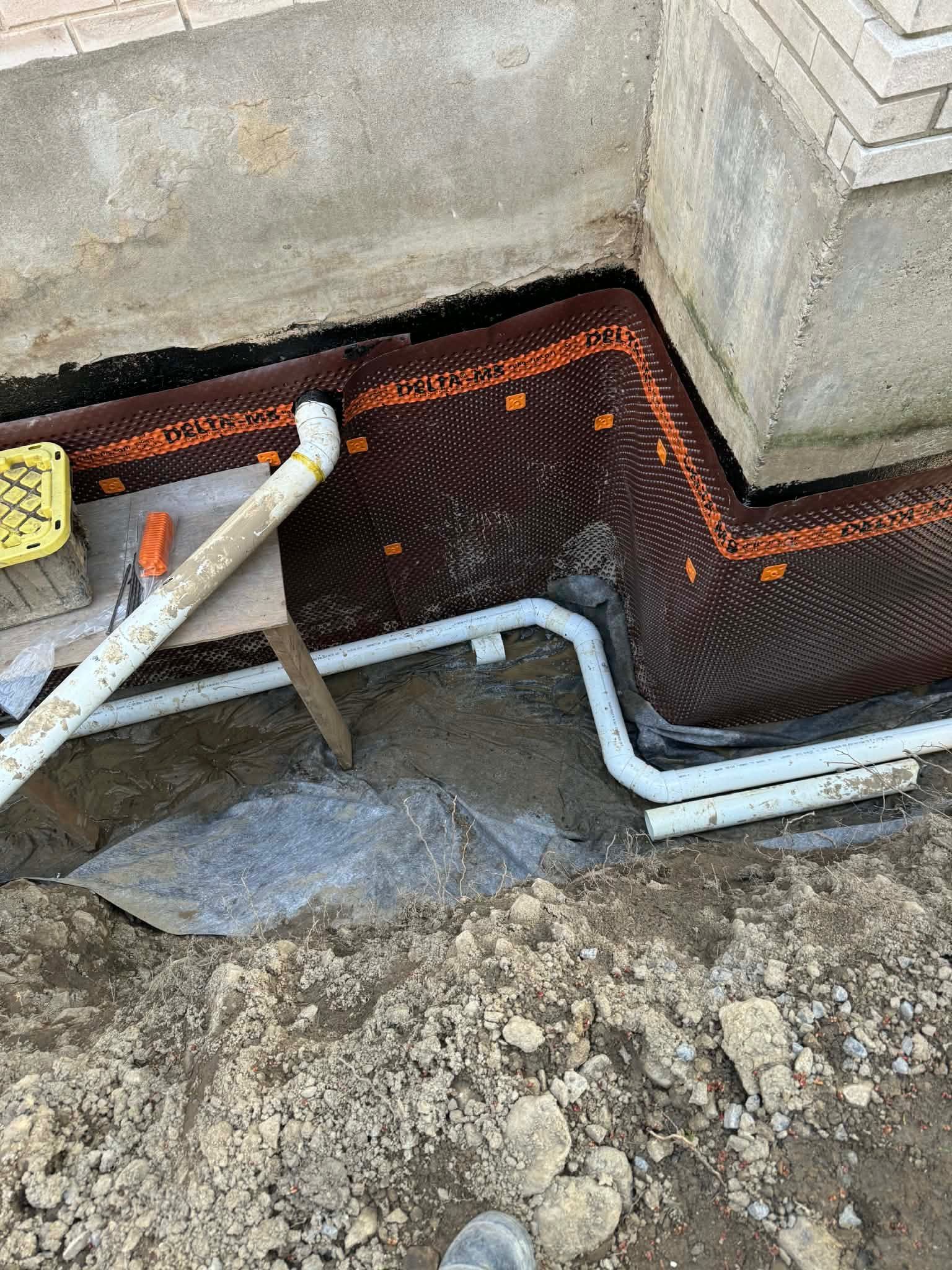 Black drainage mat and white pipe installed along a concrete foundation trench.