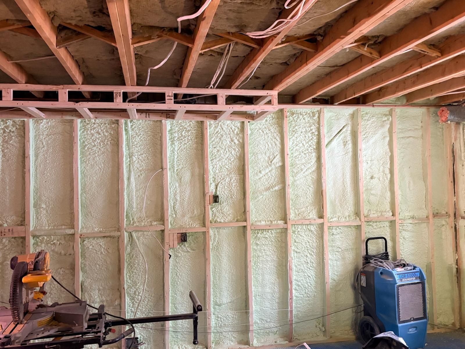 Basement wall with spray foam insulation, exposed ceiling joists, and construction tools in the foreground