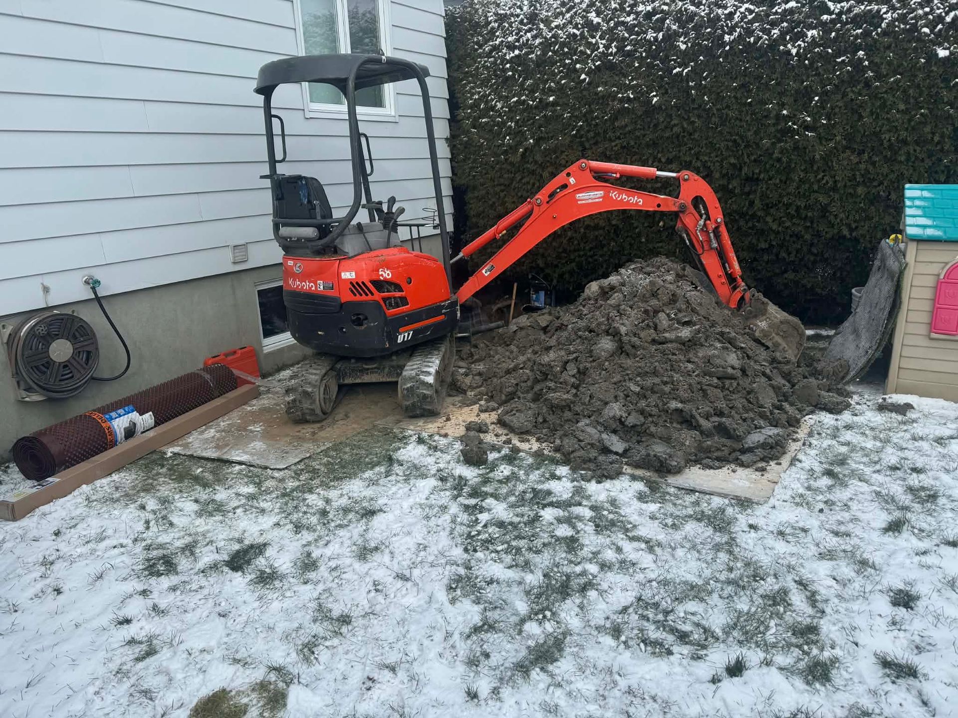 Small orange excavator digging a mound of dirt beside a house on a snowy yard