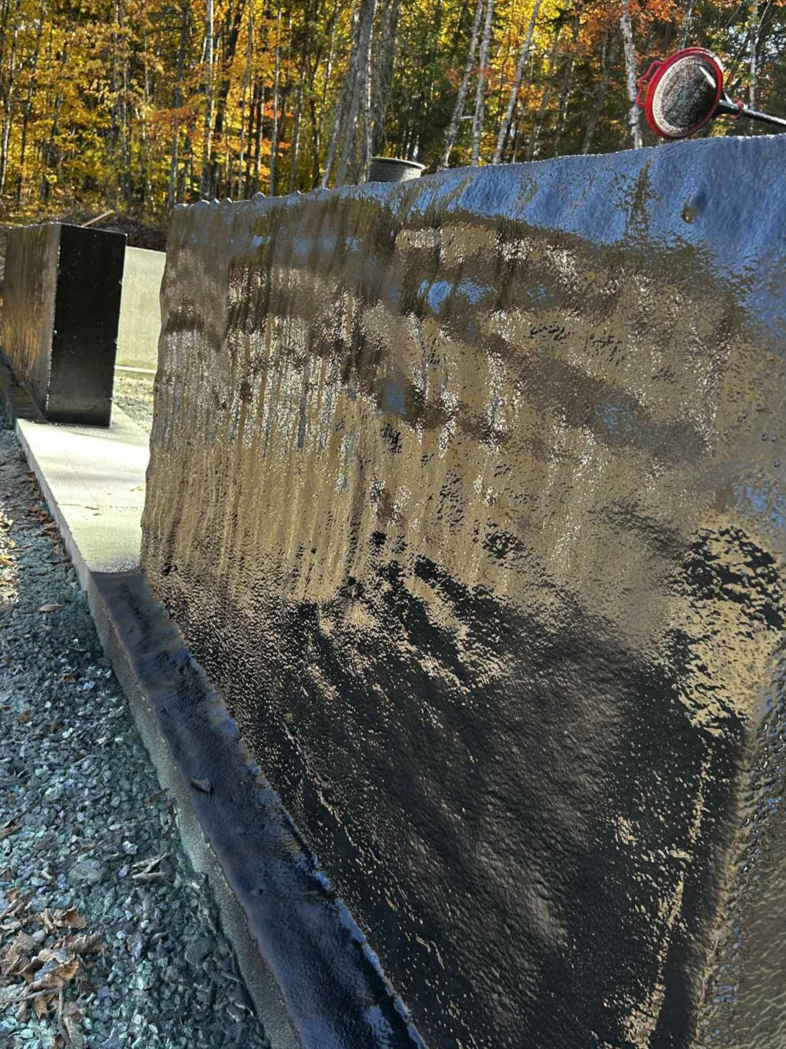 Dark concrete retaining wall beside gravel path, with trees in autumn and a rock on top.
