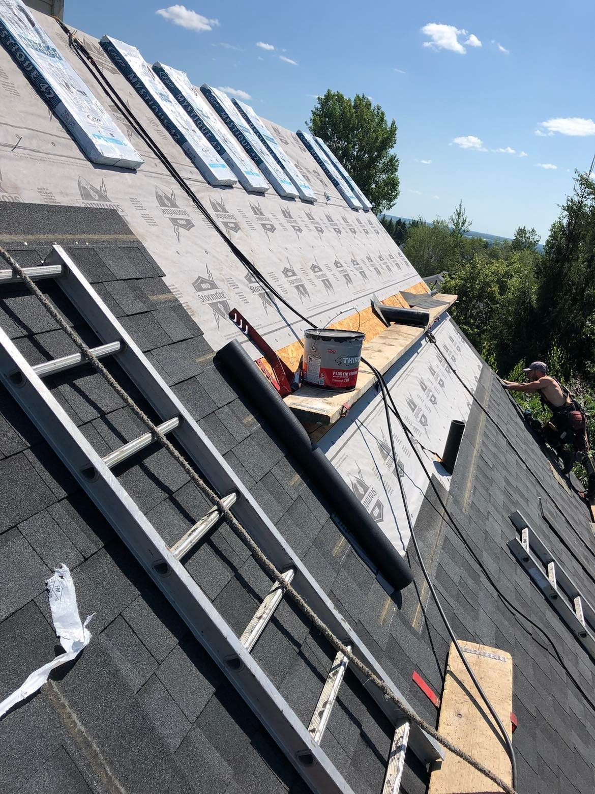 Roof under construction with black shingles, ladder, lumber, and tools on a steep slope.