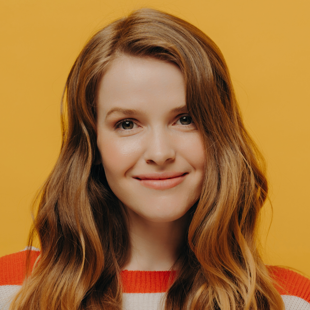 Woman with auburn hair smiles at the camera against a yellow backdrop, wearing a white and orange striped sweater.