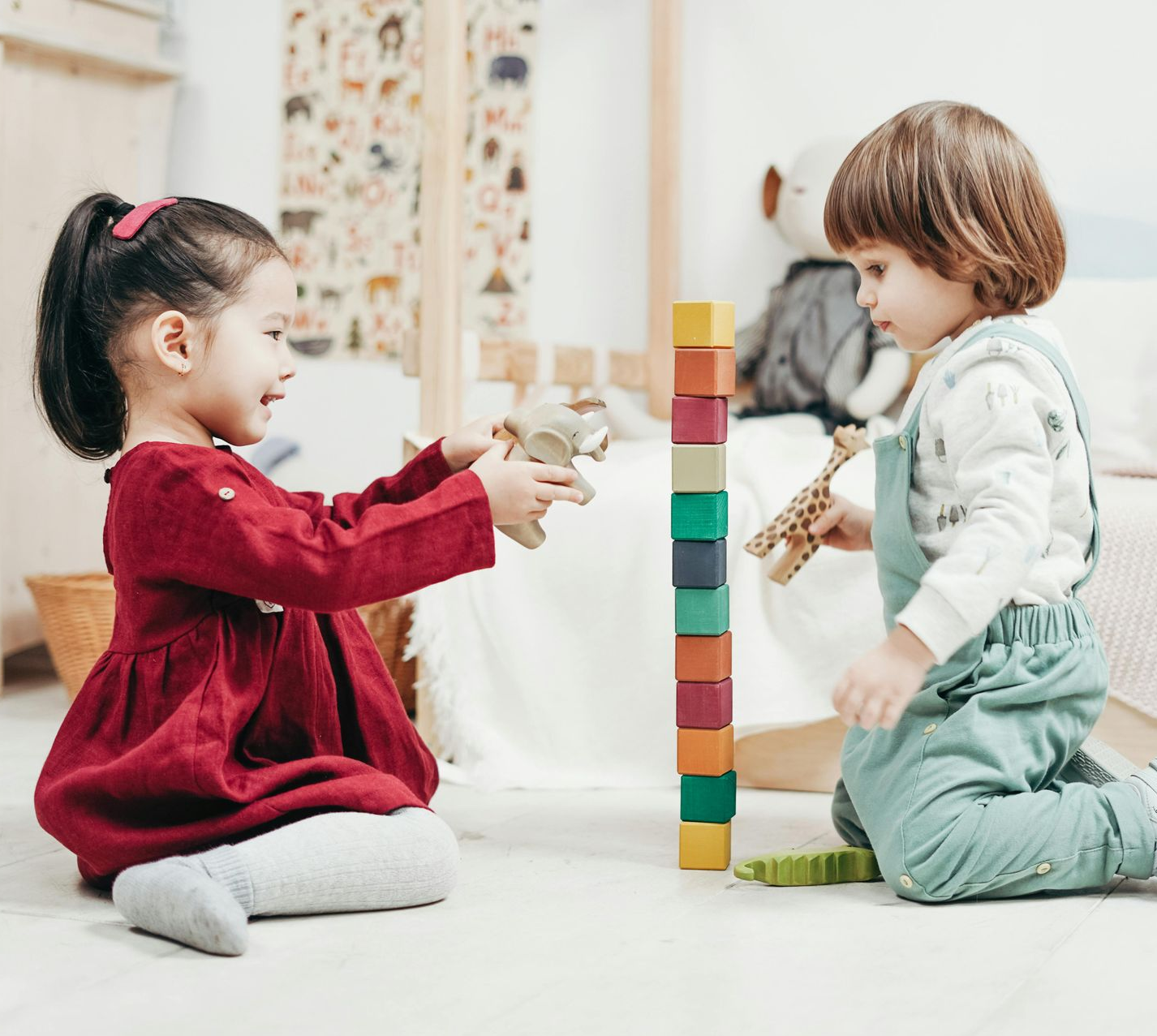 Two children play together on a floor, building a colorful block tower and holding toy animals in a bright room.