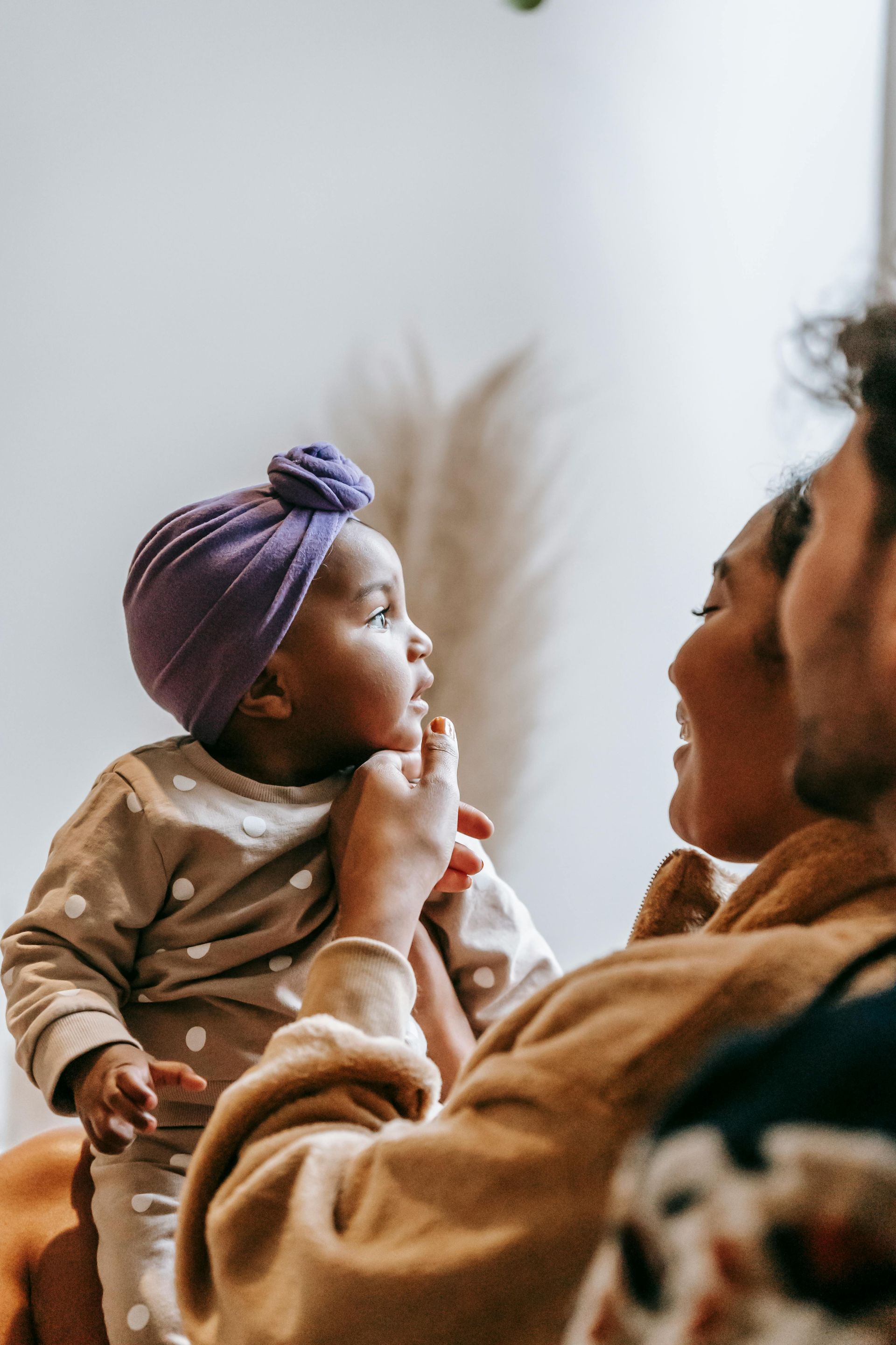 A baby with a purple head wrap gazes up at smiling parents; setting is indoors.