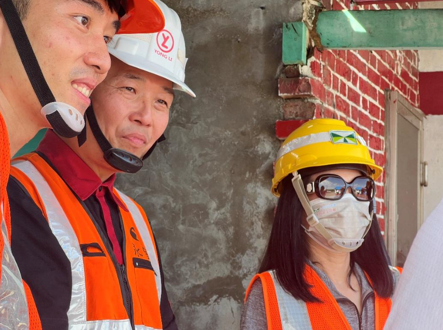 三個戴著安全帽、穿著安全背心的人站在建築工地的一面紅磚牆旁。
