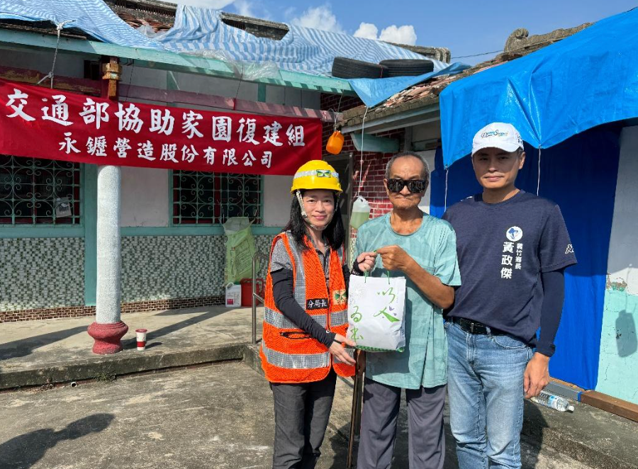 三個人站在一棟建築物前,面前舉著橫幅,其中一人穿著安全背心和頭盔,手裡拿著一個禮品袋。