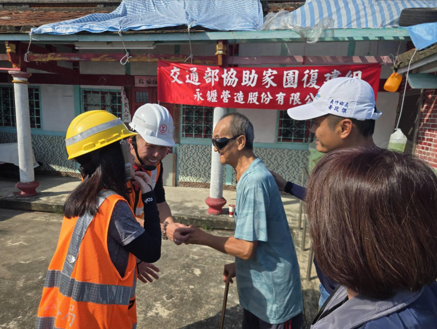 在工地現場,身穿高能見度背心、頭戴安全帽的工人與一位老人在紅色橫幅前握手。