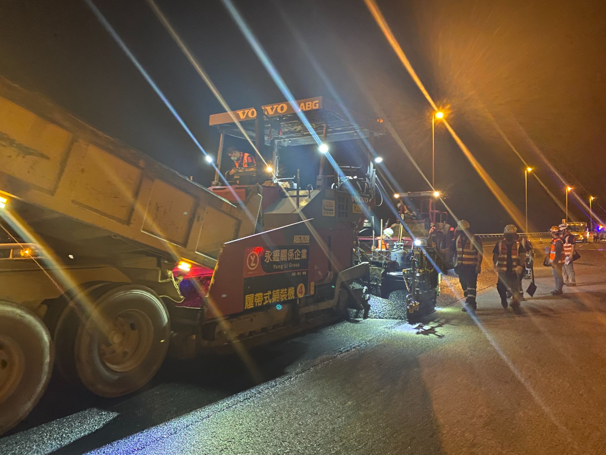 Nighttime roadwork with a large dump truck and workers beside an asphalt paving machine under streetlights.