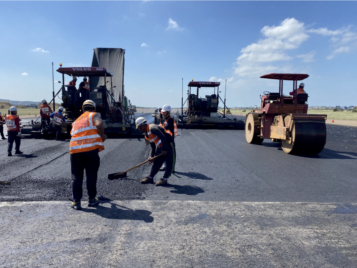 Workers operating road pavers on a highway under construction, with orange safety vests and heavy machinery.
