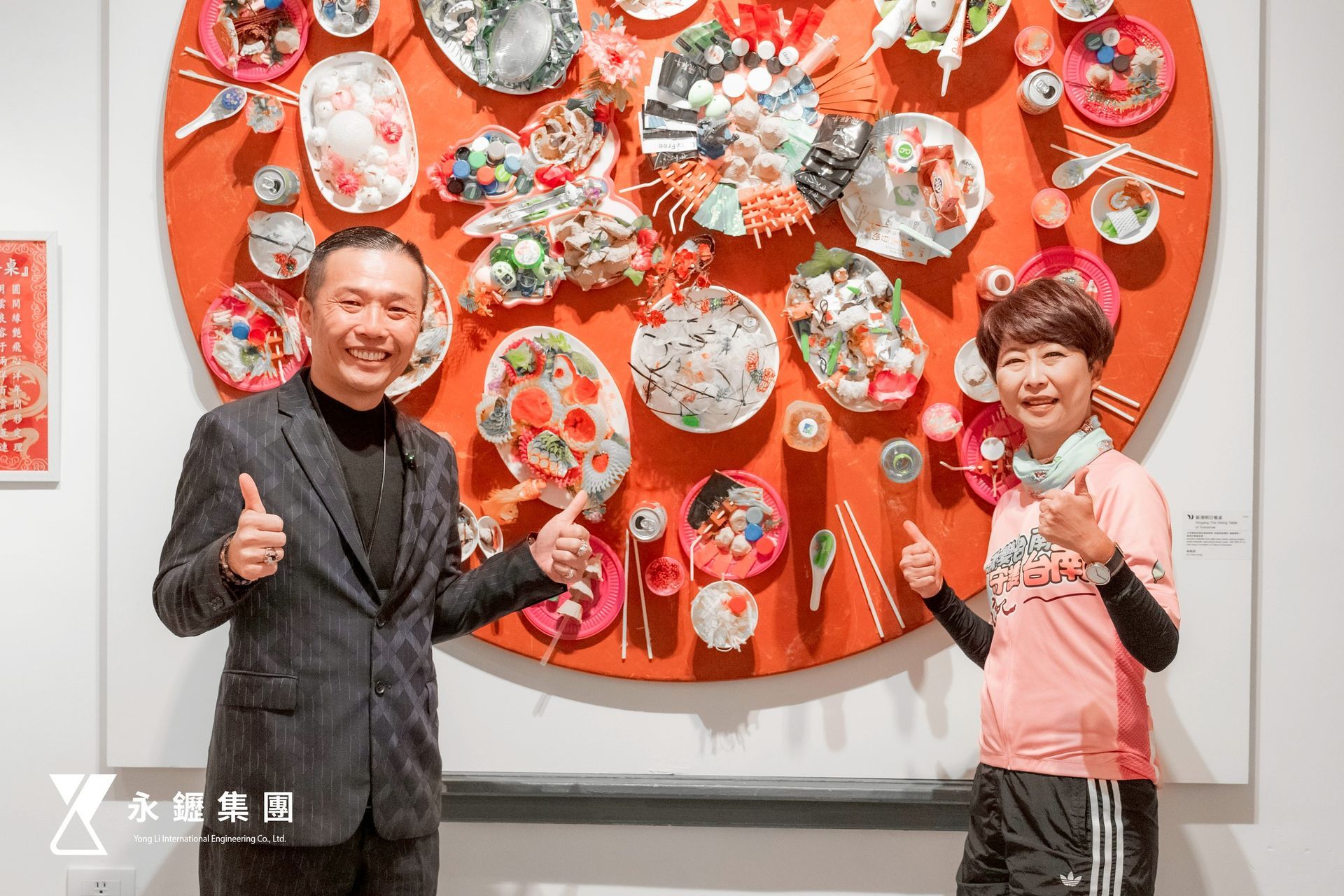 一名男子和一名身穿運動服的人站在一個巨大的圓形橙色藝術品前,兩人都豎起了大拇指。