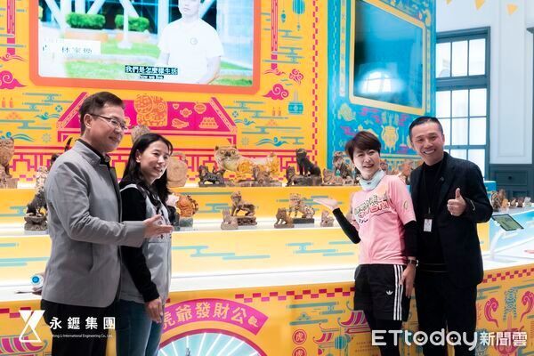四個人站在一個活動現場色彩繽紛的小動物模型前,活動現場有醒目的黃色標誌和品牌標誌。