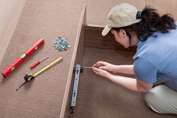 Person assembles furniture, using a screwdriver, near a level, tape measure, and screws.