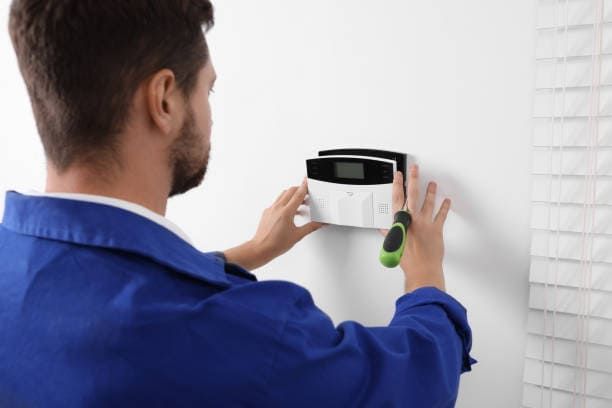 Man in blue jumpsuit installing security system panel on white wall.