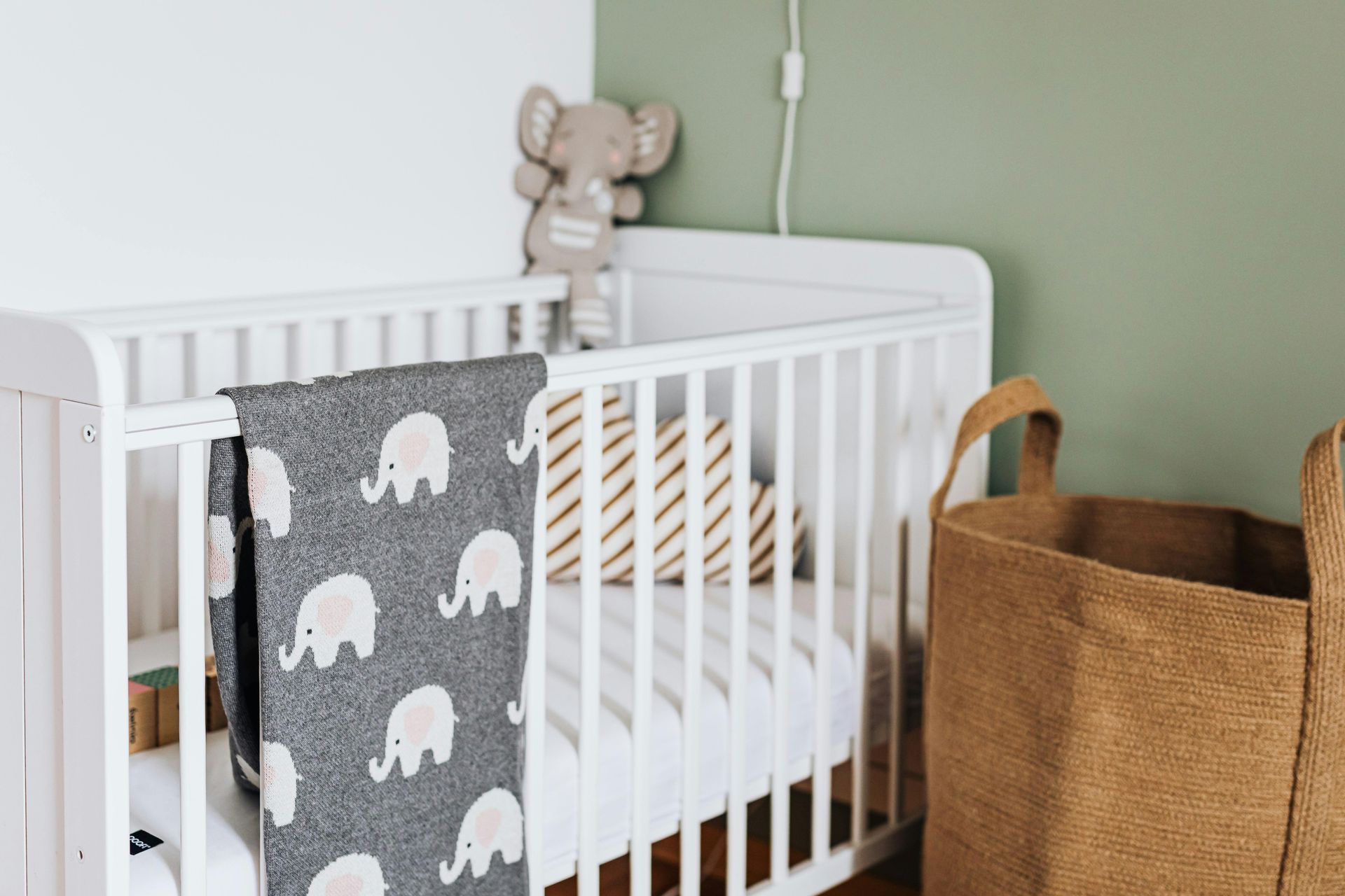 White crib with gray elephant blanket and teddy bear in a soft green nursery, beside a woven storage basket