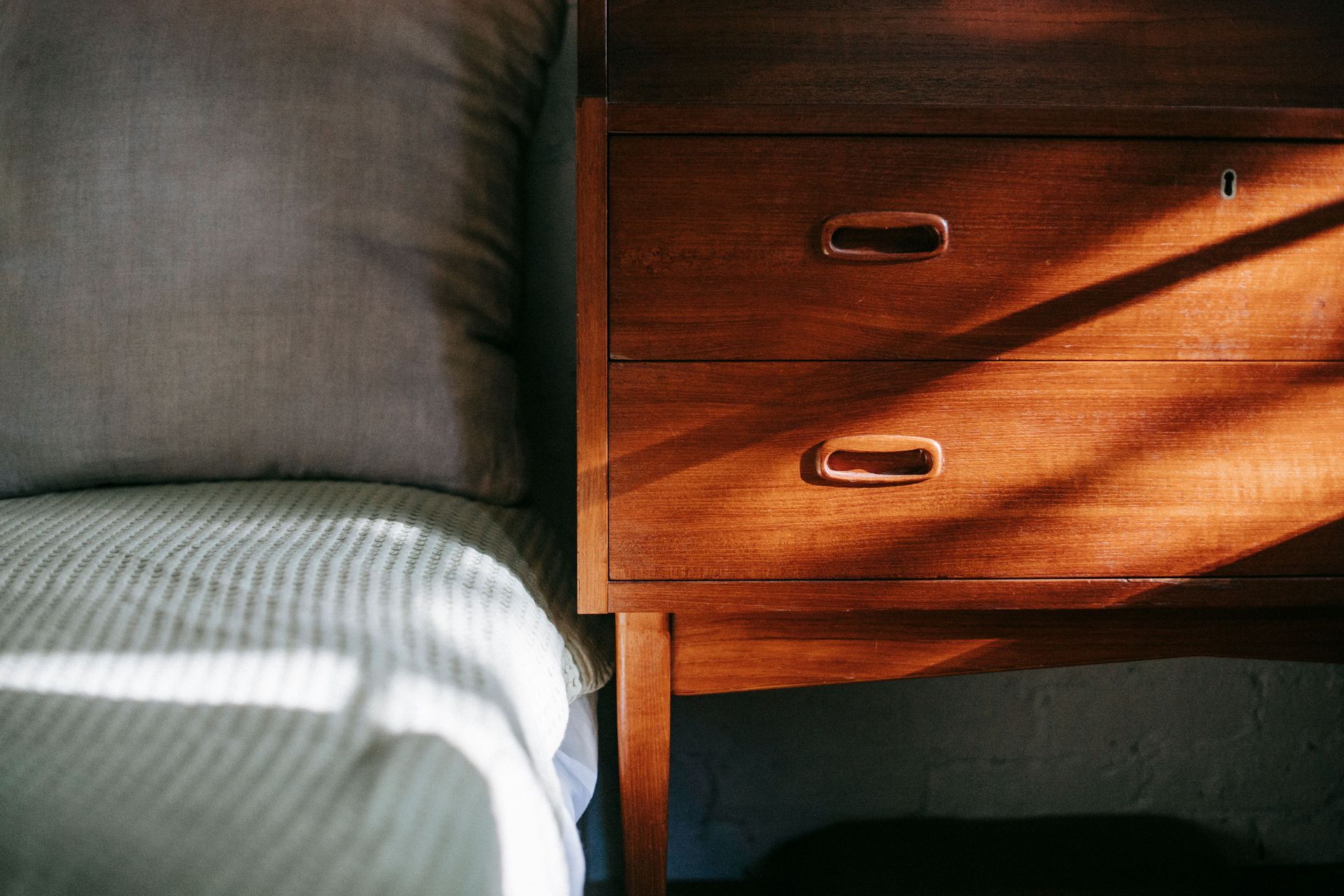 Bedside table beside a gray bed in warm sunlight, with shadows across the wooden drawers.