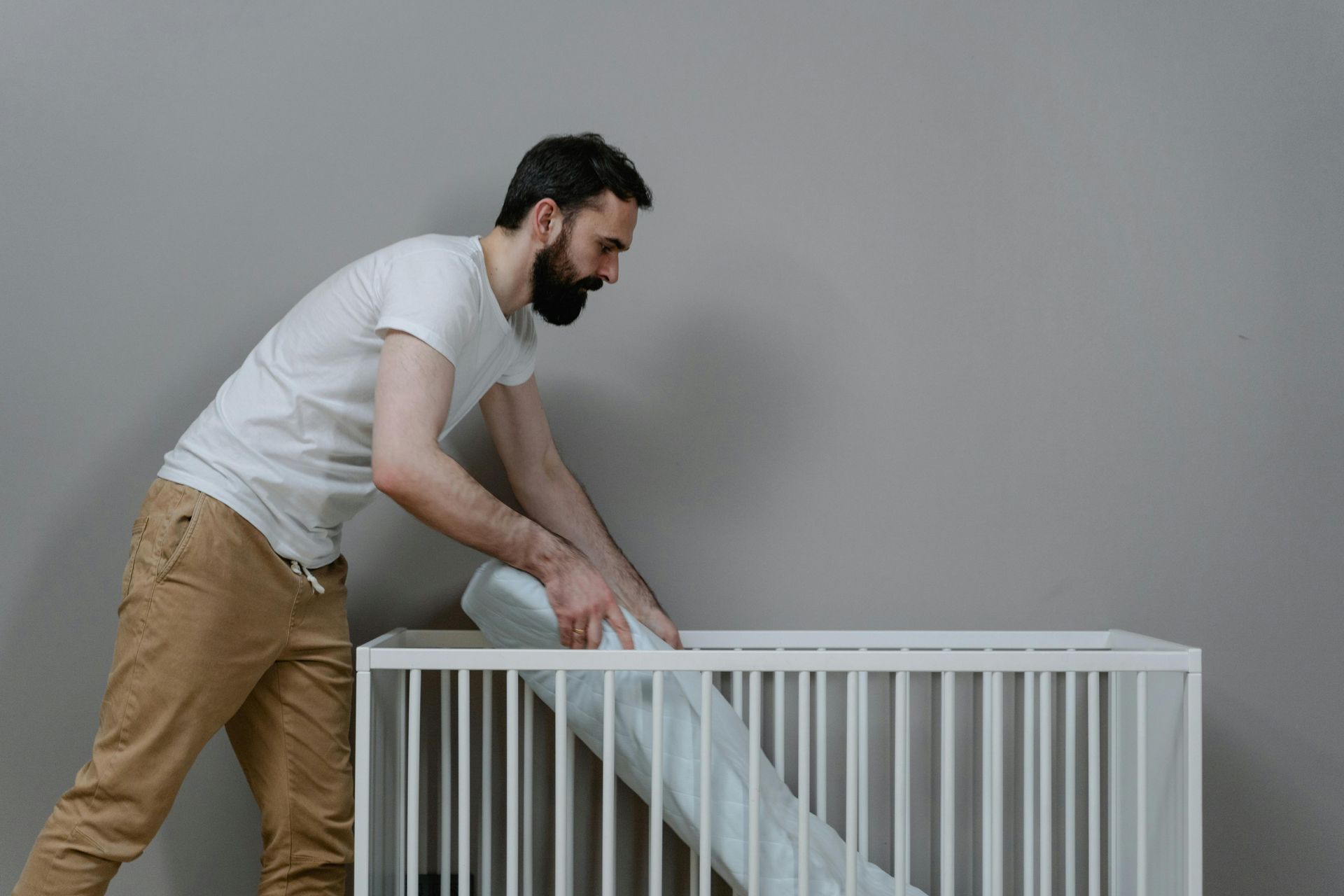 Man in white T-shirt and tan pants adjusting a blanket in a white crib or bed against a gray wall