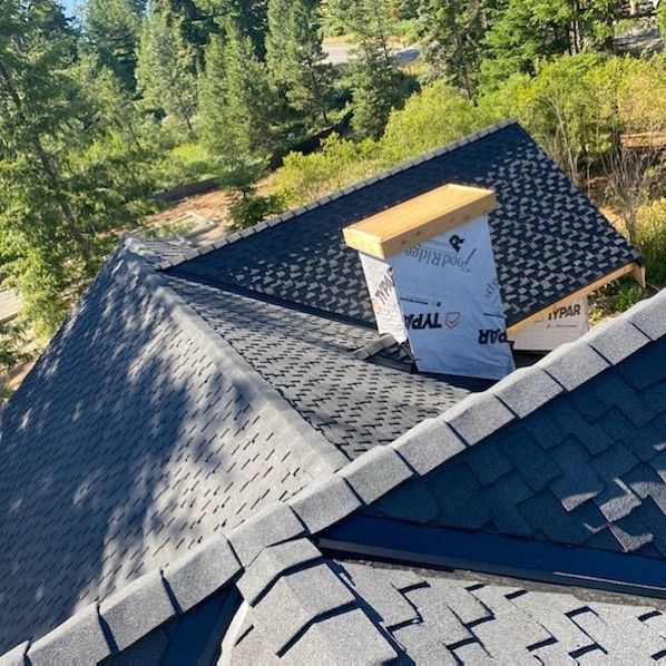 Roof with dark shingles and a chimney under construction in a wooded area.