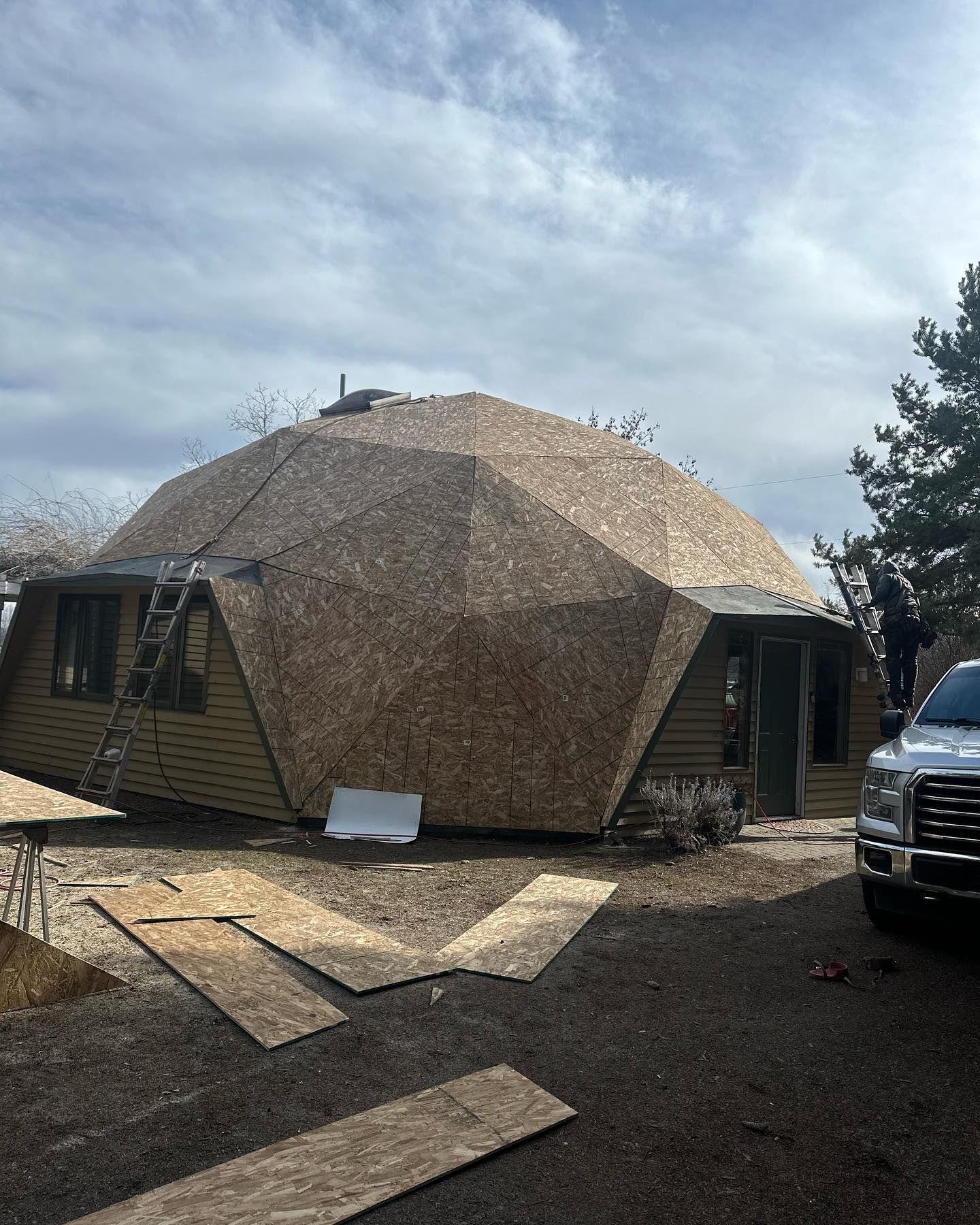 A dome-shaped house under construction, covered with wooden panels. A person stands on a truck bed.