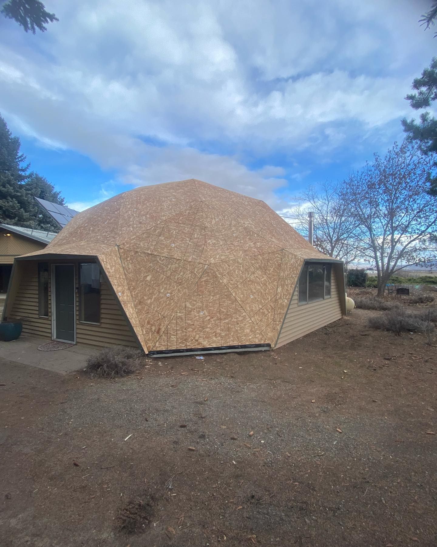 Geodesic dome house with plywood roof and tan siding under cloudy sky.