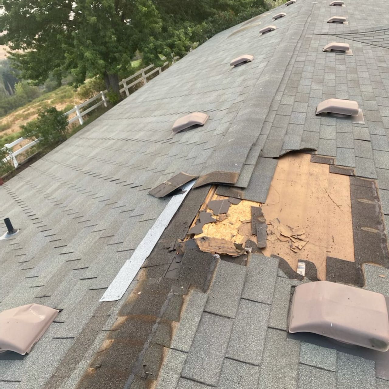 Damaged asphalt shingle roof with missing shingles exposing wooden decking. Several vents are visible.