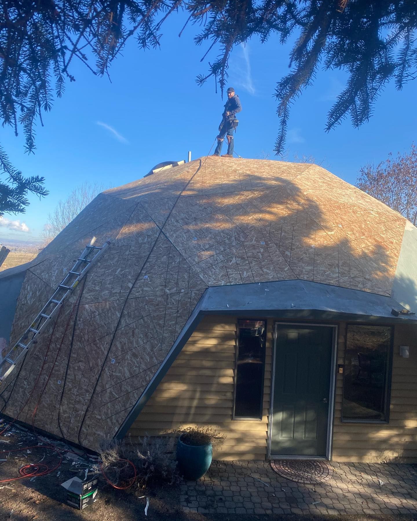 Man on a geodesic dome roof, installing roofing panels. A-frame structure with a ladder. Blue sky backdrop.