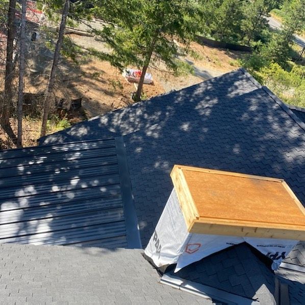 Roof with dark shingles and a wooden box-like structure, trees and a hillside in the background.