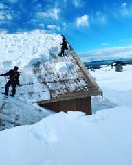 Two people clearing snow from a brown roof under a bright blue sky.