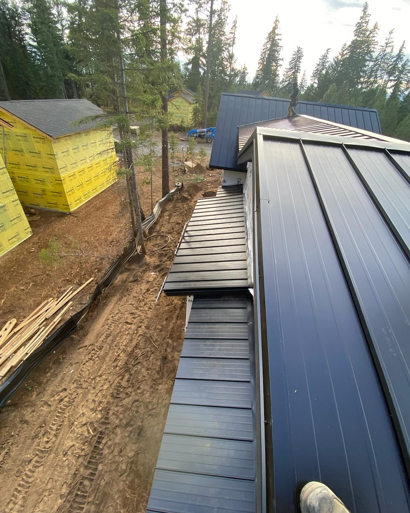 View of a building's roof under construction; dark metal panels and black trim. Yellow-wrapped structure and trees in background.