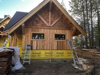 Wooden building under construction, brown siding, two black windows, ladders, forest background.