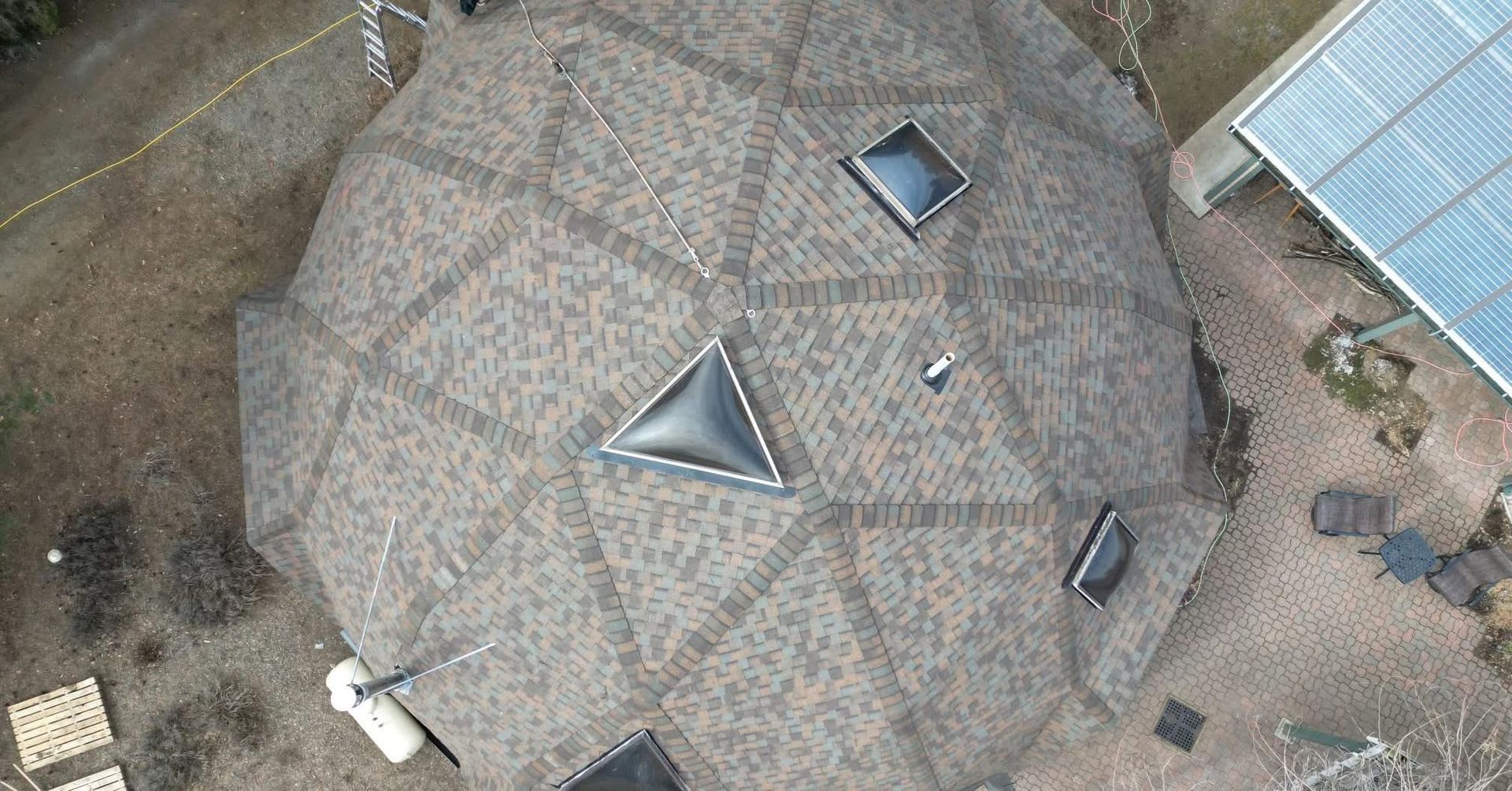 Aerial view of a geodesic dome roof with triangular and rectangular skylights. Brown shingles. Solar panels are visible.