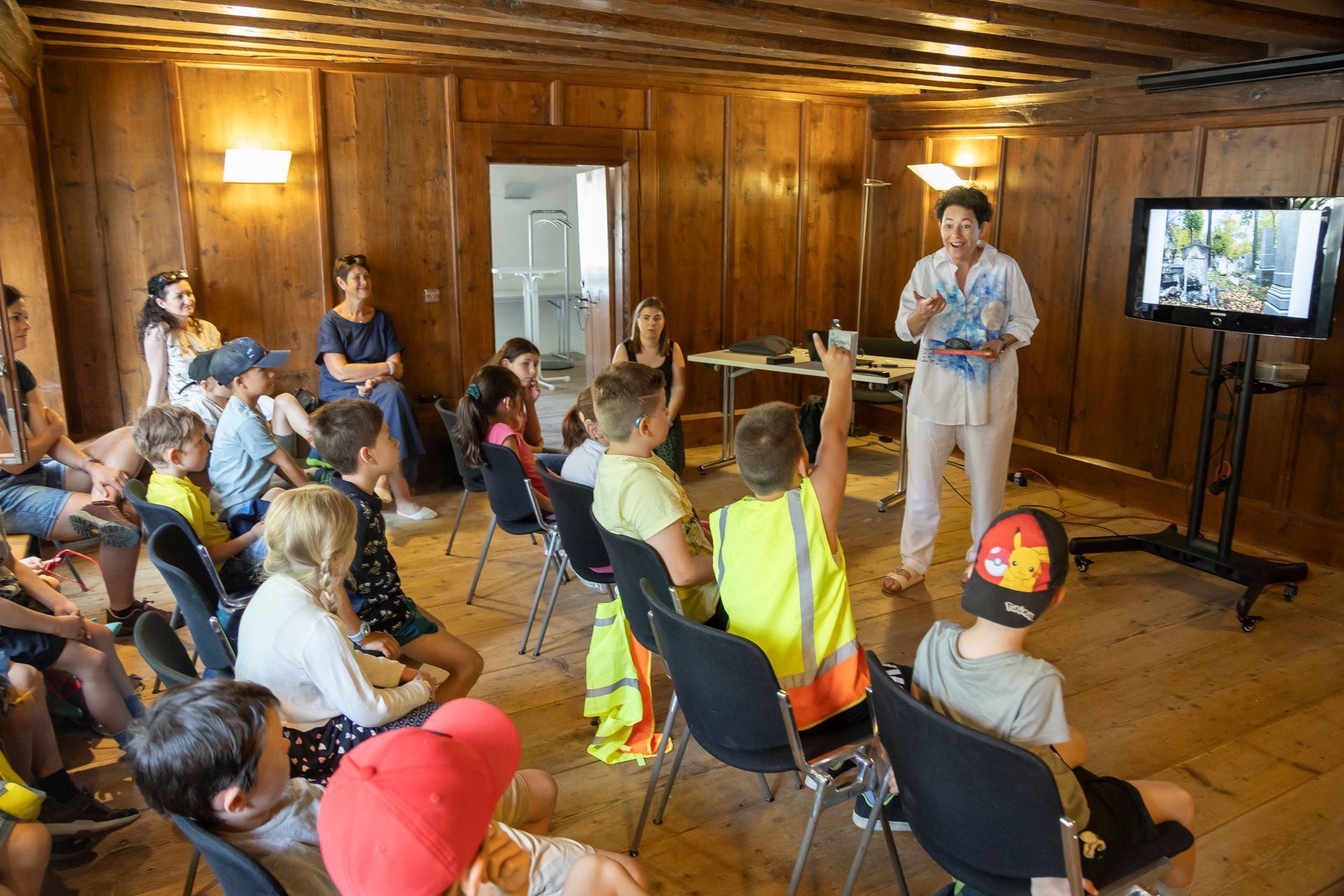 Eine Gruppe Kinder sitzt in einem Raum, vor ihnen steht eine Frau.