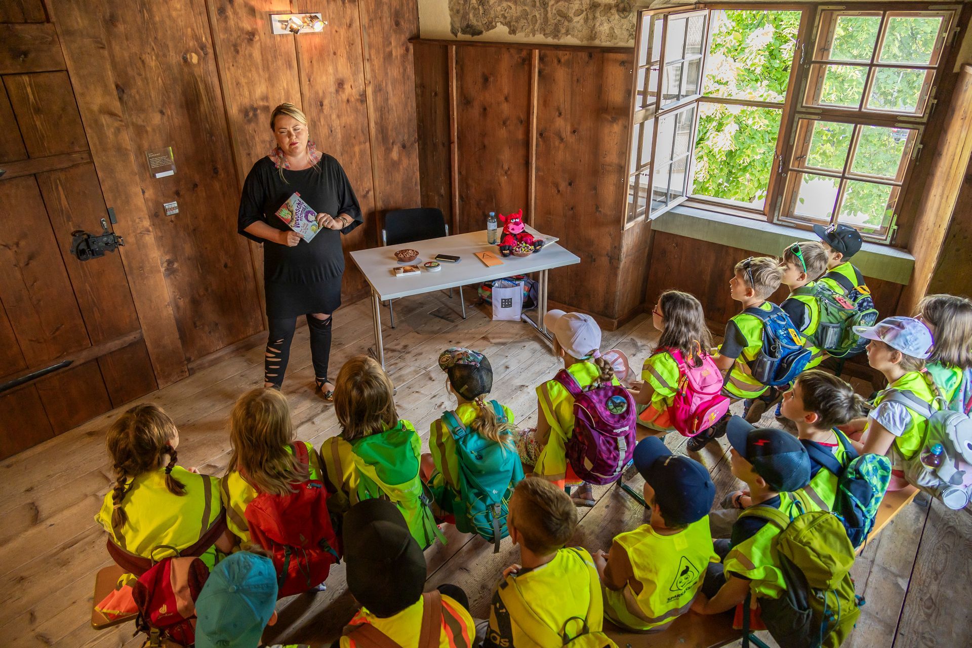 Eine Frau steht vor einer Gruppe von Kindern in einem Raum mit eimem Buch.