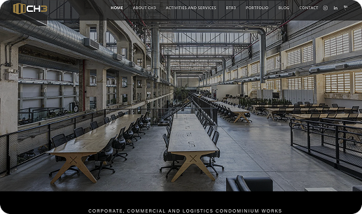 Sala comprida em estilo industrial com fileiras de mesas e cadeiras, possivelmente um espaço de trabalho ou refeitório.