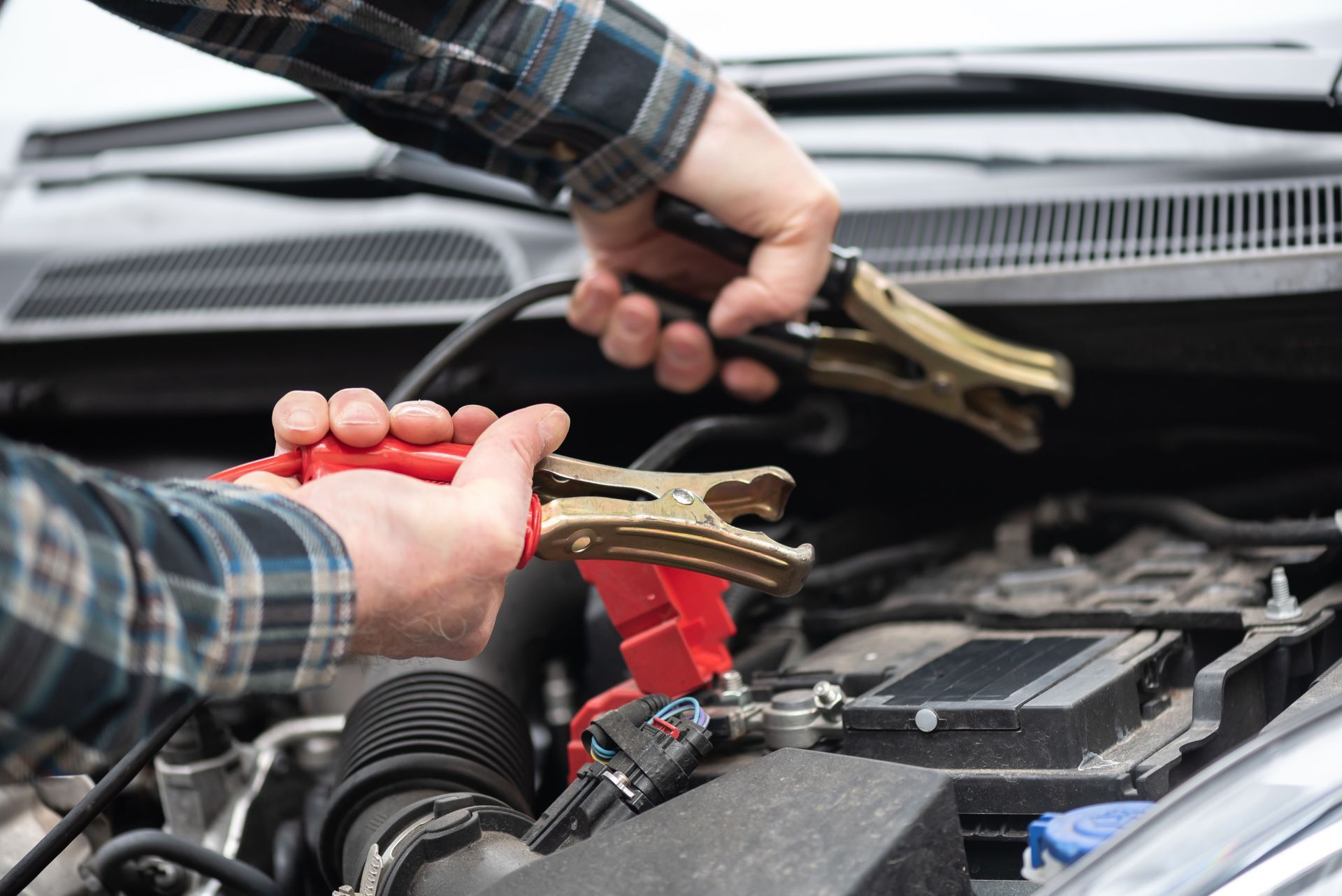 Hands in plaid sleeves holding gold-toned jumper cable clamps over a car battery engine compartment.