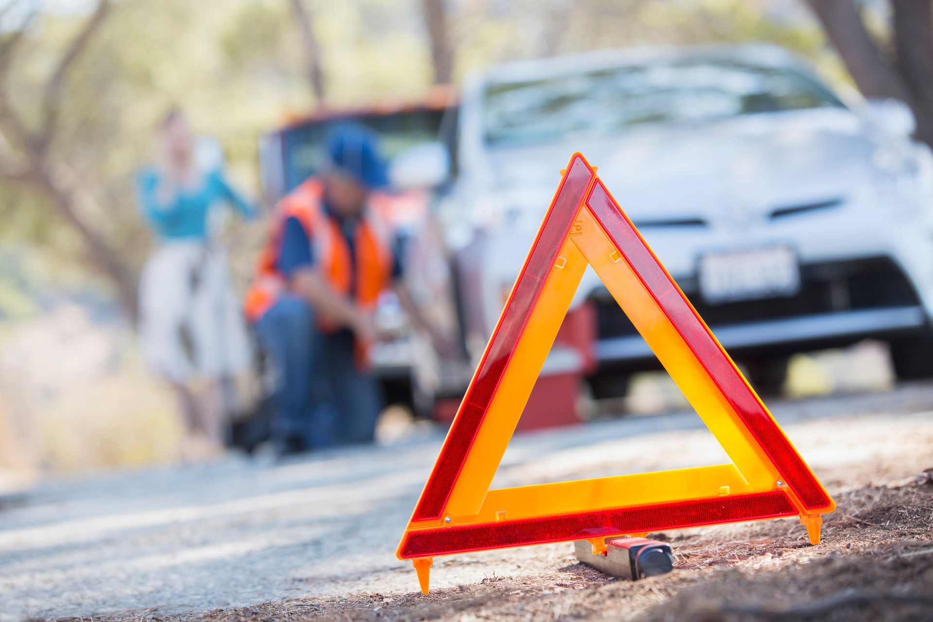 A bright red-and-orange warning triangle sits on a roadside in the foreground, with blurred people and a car behind.