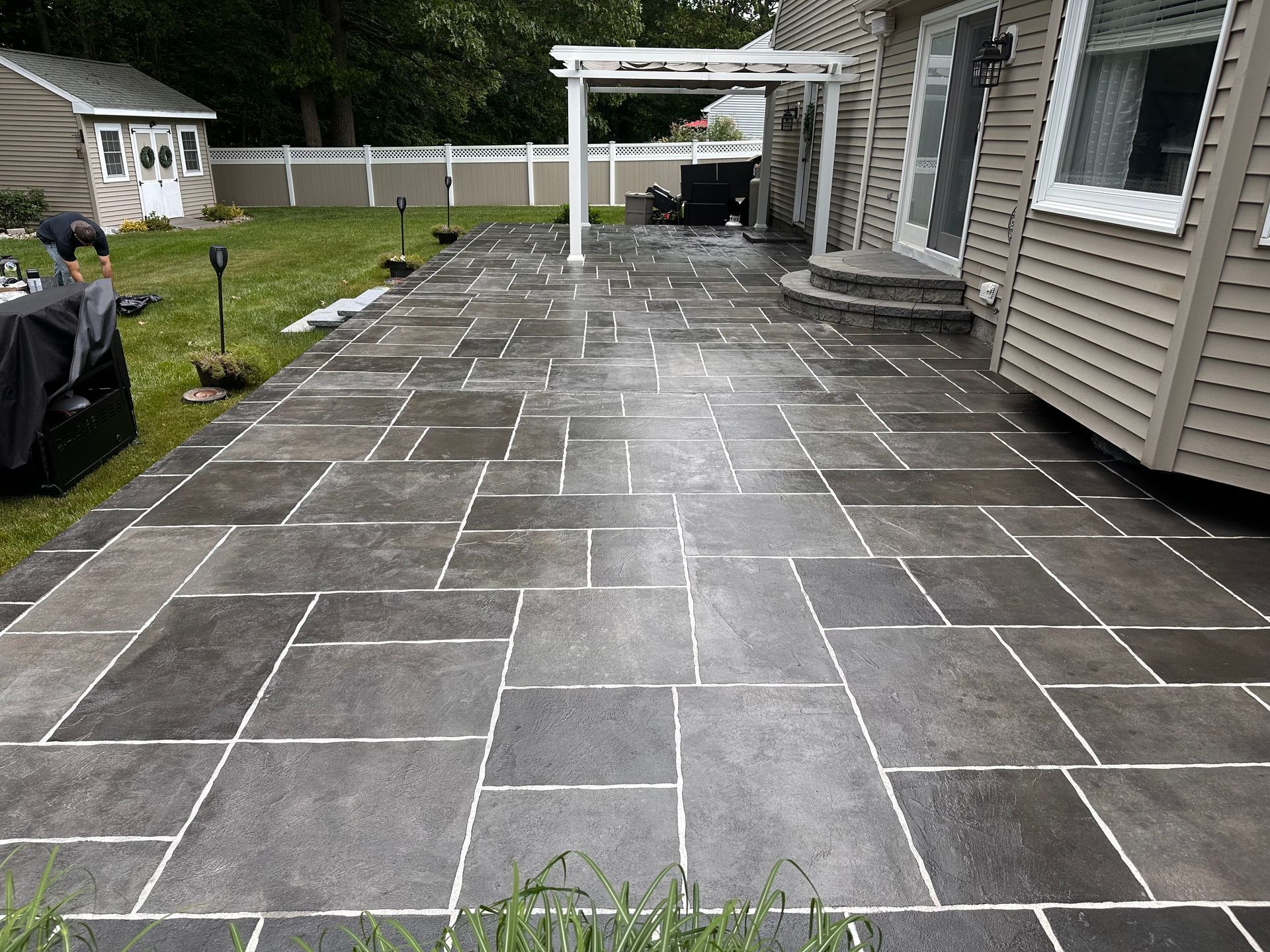Wet stone patio beside a house, with a pergola and fenced yard in the background.