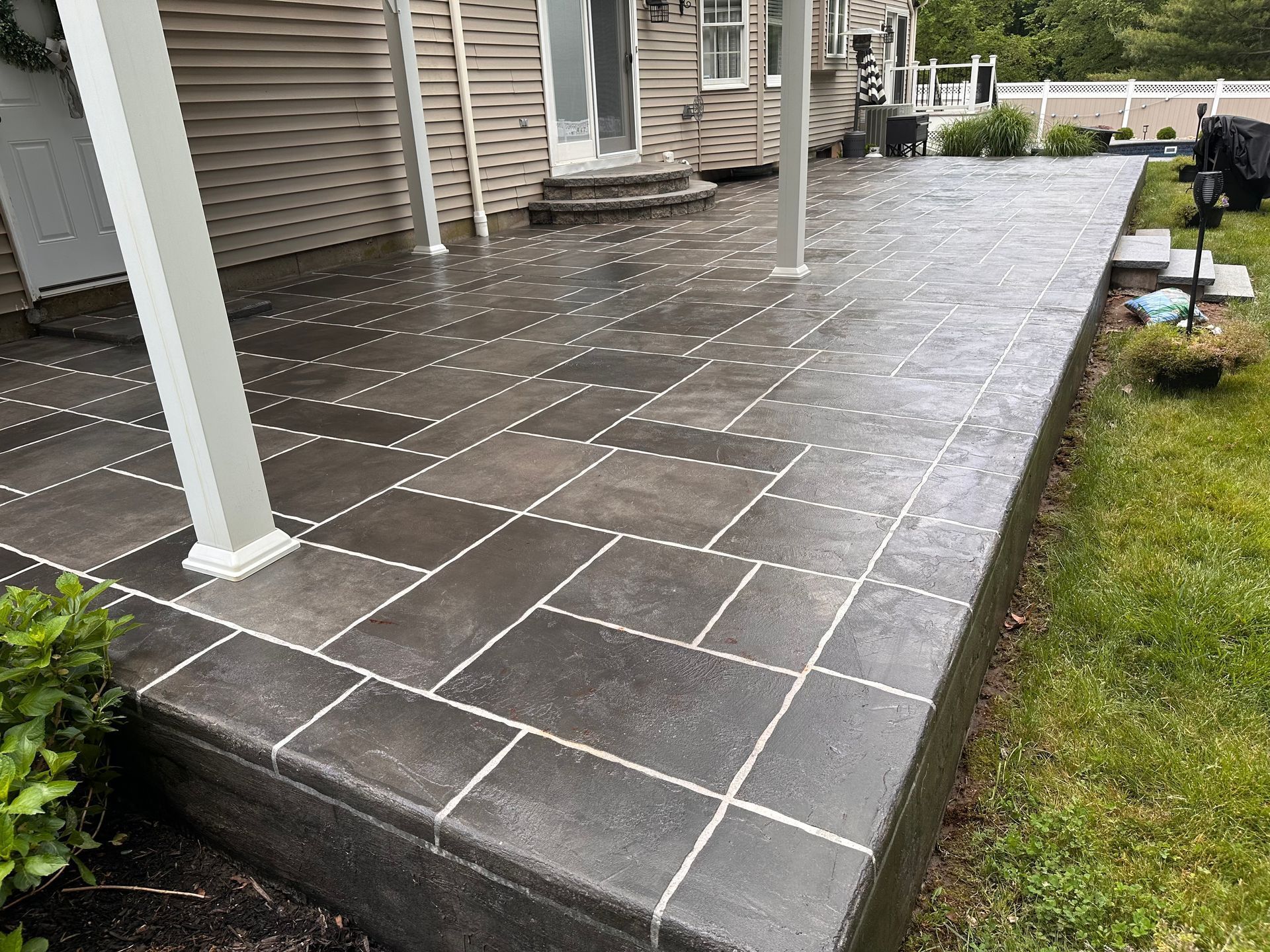 Covered patio with dark tile flooring, white columns, and a wet walkway beside a grassy yard