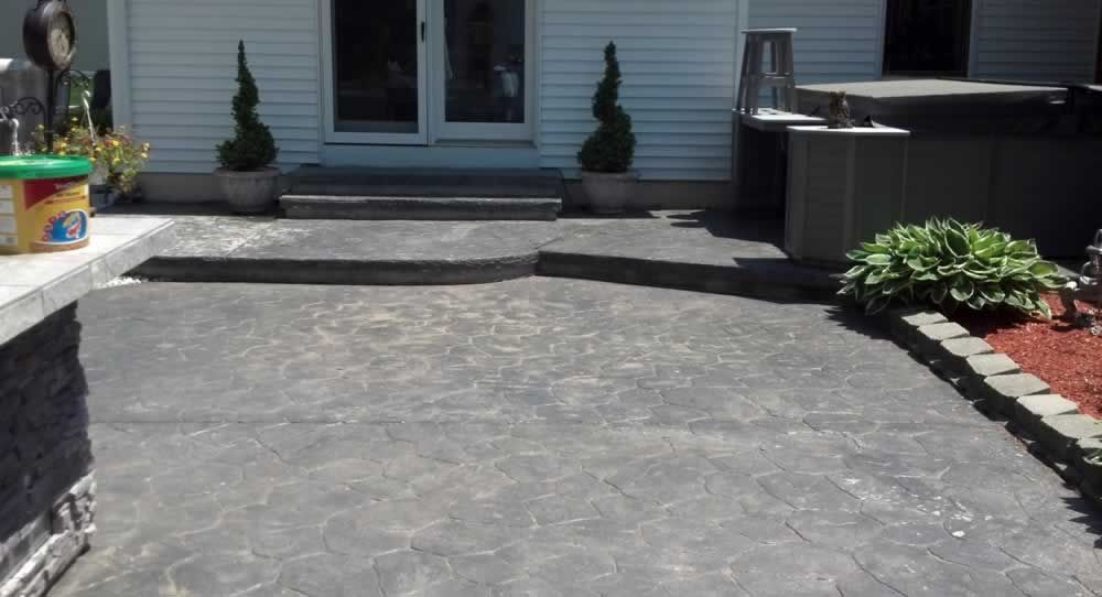 Backyard patio with a gray stamped concrete surface leading to a house entrance, bordered by plants and brick edging