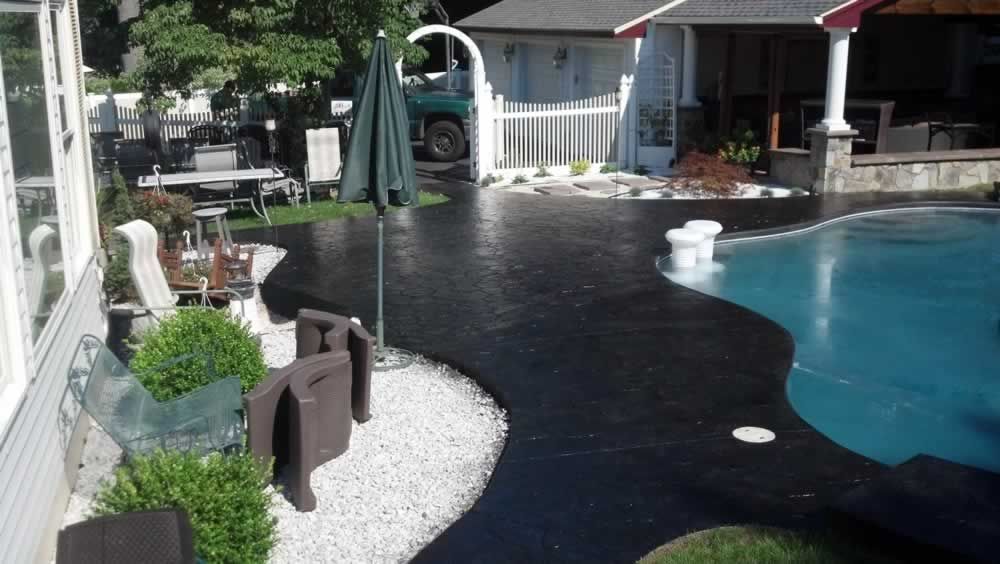 Backyard pool with black patio, lounge chairs, umbrella, and house in the background