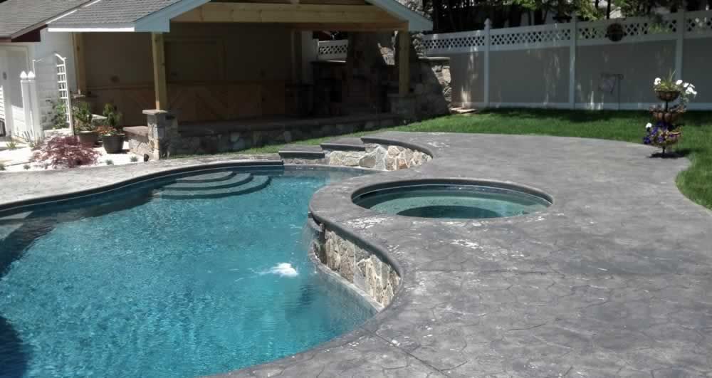 Backyard pool with a stone spa, curved concrete deck, and covered patio beside a fence and lawn