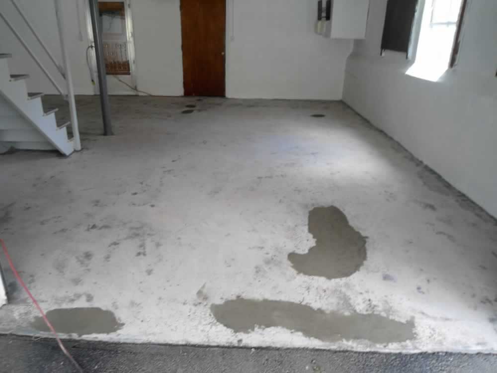 Empty unfinished room with gray concrete floor, white walls, stairs on the left, and windows on the right.