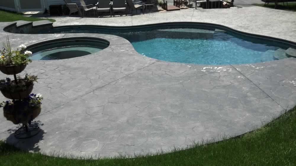 Curved concrete pool deck beside a blue swimming pool and round hot tub, with grass edging the patio