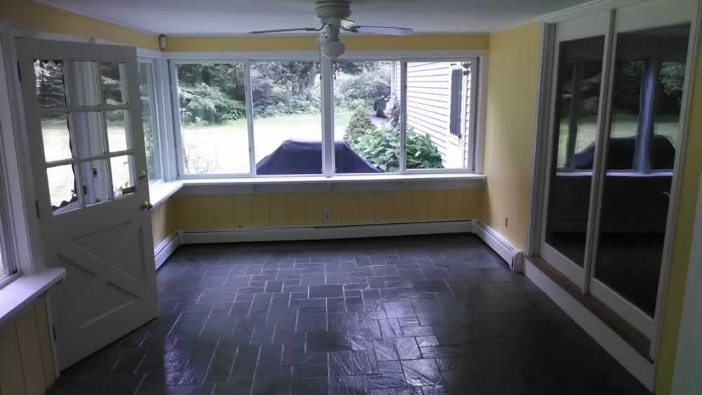 Empty enclosed porch with dark tile floor, yellow walls, and large windows overlooking greenery.