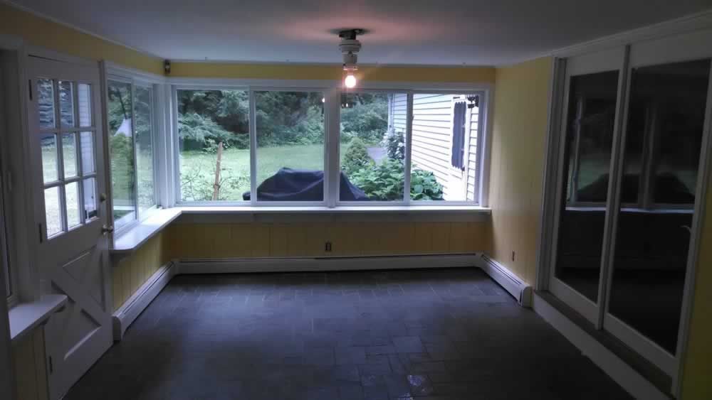 Empty enclosed porch with large windows and gray carpet, overlooking a backyard.