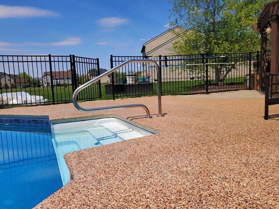 Backyard pool with tan pebble deck, metal handrail, and fenced grassy area under a sunny sky