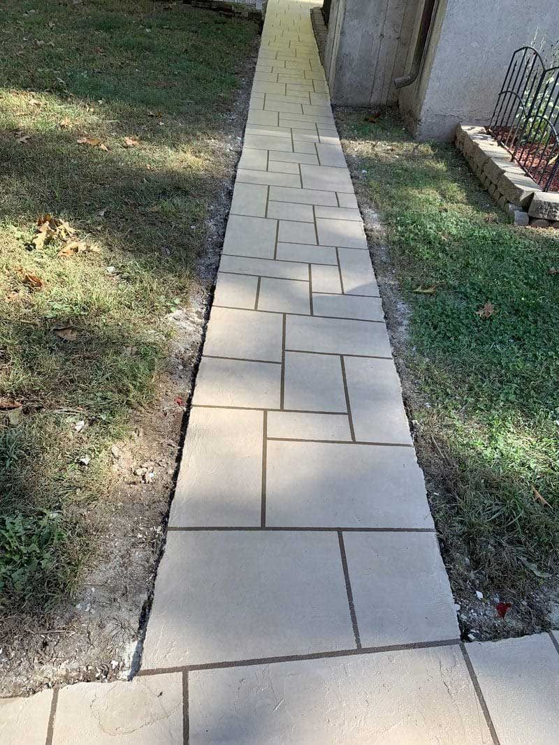 Narrow concrete garden walkway between grass and a house wall.