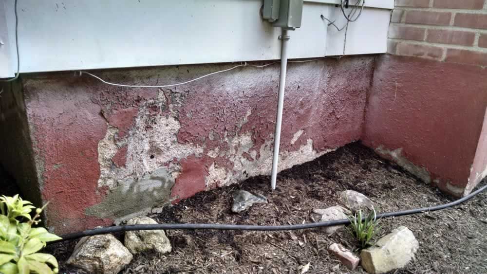 Peeling red foundation wall beneath house, with exposed damage and pipes along the base.