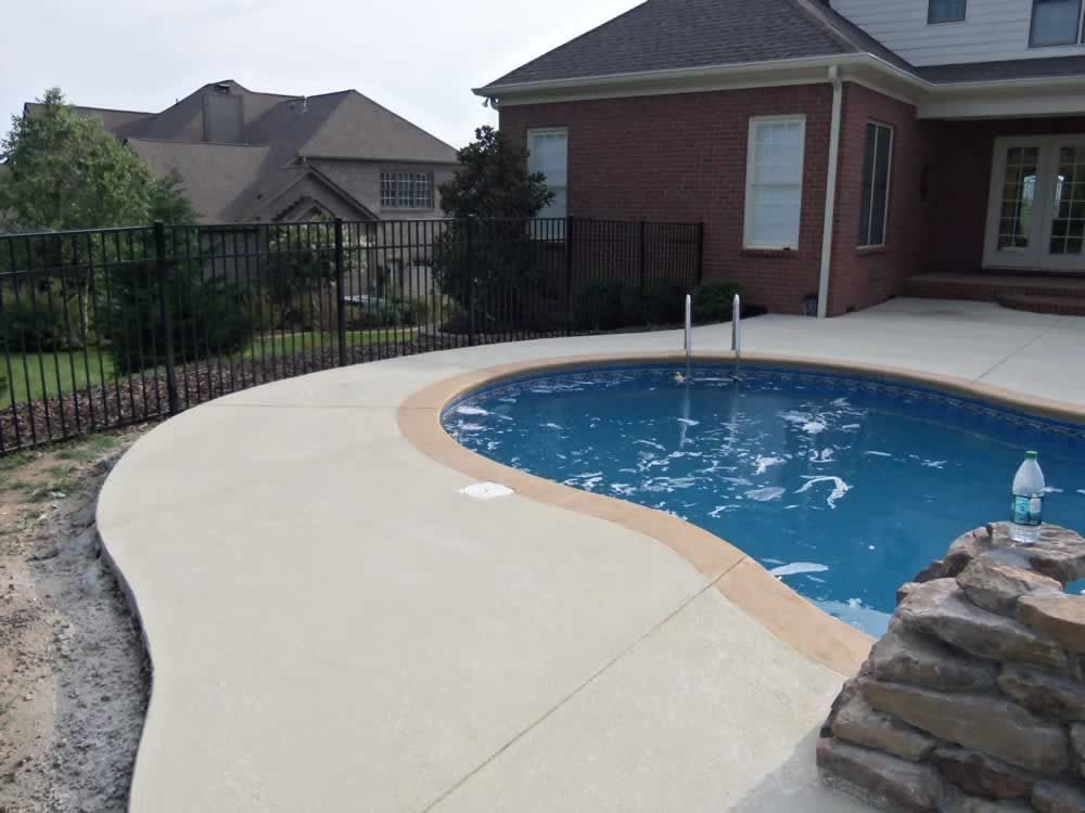 Backyard swimming pool with curved concrete deck beside a brick house and fence