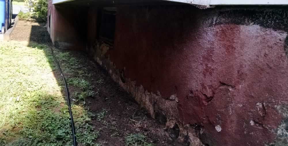 Damp red wall under a building with dark mold stains and a narrow grassy strip beside it